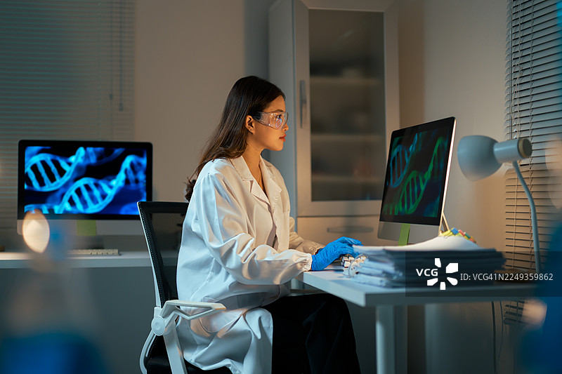 年轻的女性科学家穿着实验服，戴着安全眼镜，在研究实验室的电脑上工作，分析DNA数据图片素材