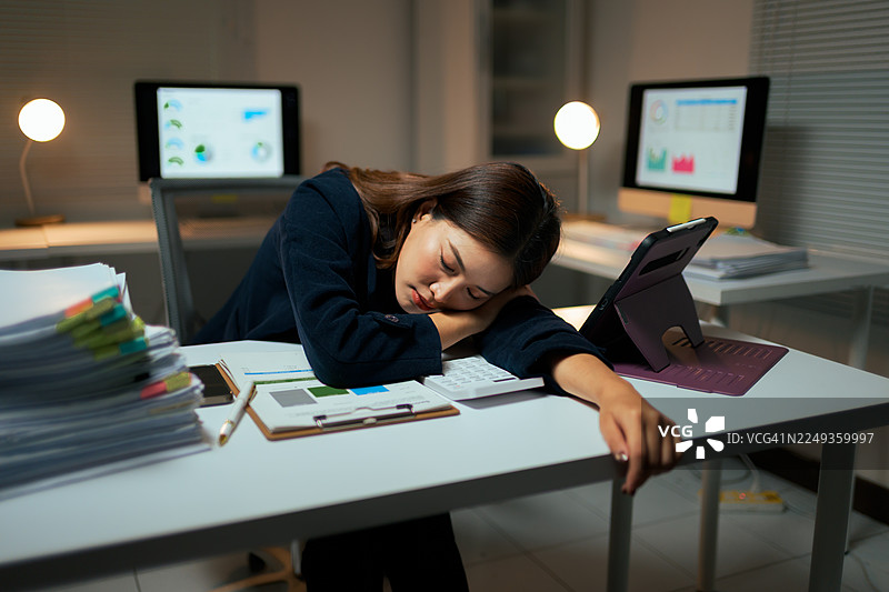 疲惫的女商人在昏暗的办公室里睡在办公桌上，周围堆满了文件和电脑，她正经历着职业倦怠和压力。图片素材