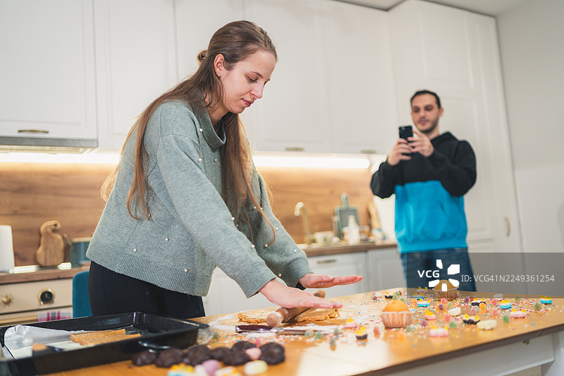 情侣在厨房——男人给触摸姜饼面团的女人拍照图片素材