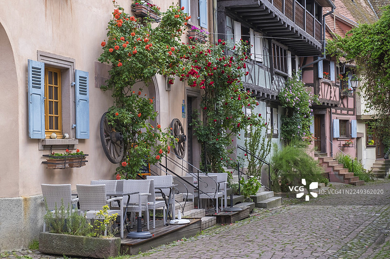 狭窄巷道里的街角咖啡馆，两旁是带有植物和红色鲜花的传统房屋，埃吉桑，法国最美乡村之一，上莱茵省，阿尔萨斯，法国图片素材