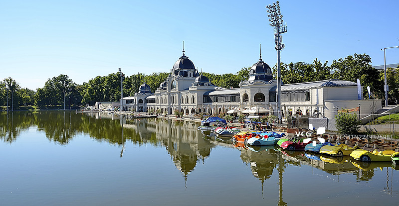 色彩鲜艳的脚踏船排在湖岸边，背景是宏伟的建筑和茂密的树木，倒映在布达佩斯城市公园平静的水面上。图片素材