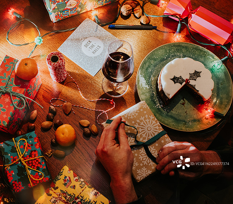 一个男人在餐桌旁打开圣诞礼物，周围摆满了节日食品和物品，如礼品、饼干和彩灯。图片素材