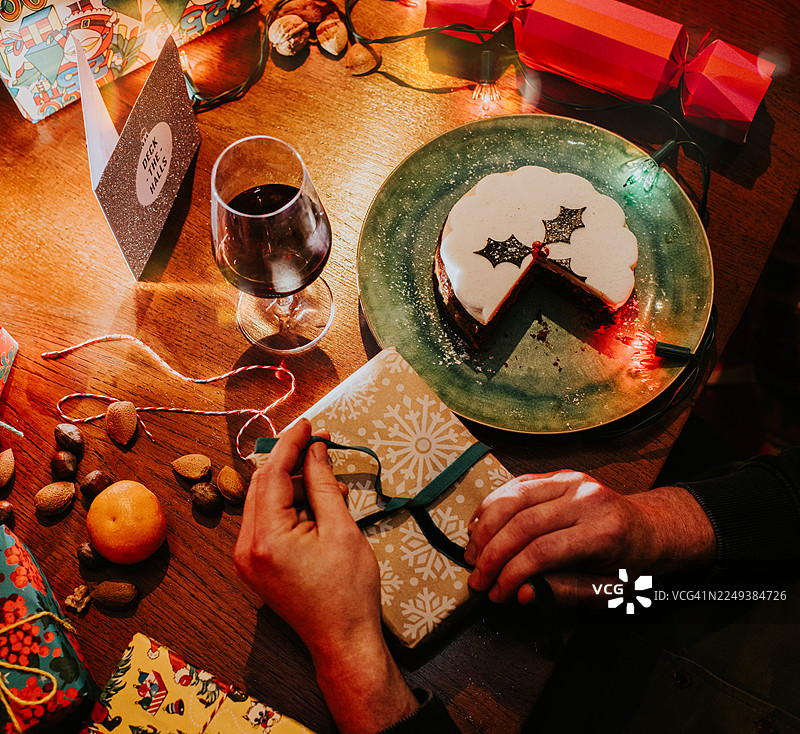 一个男人在餐桌旁打开圣诞礼物，周围摆满了节日食品和装饰品，如礼物、节日拉炮和彩灯。图片素材