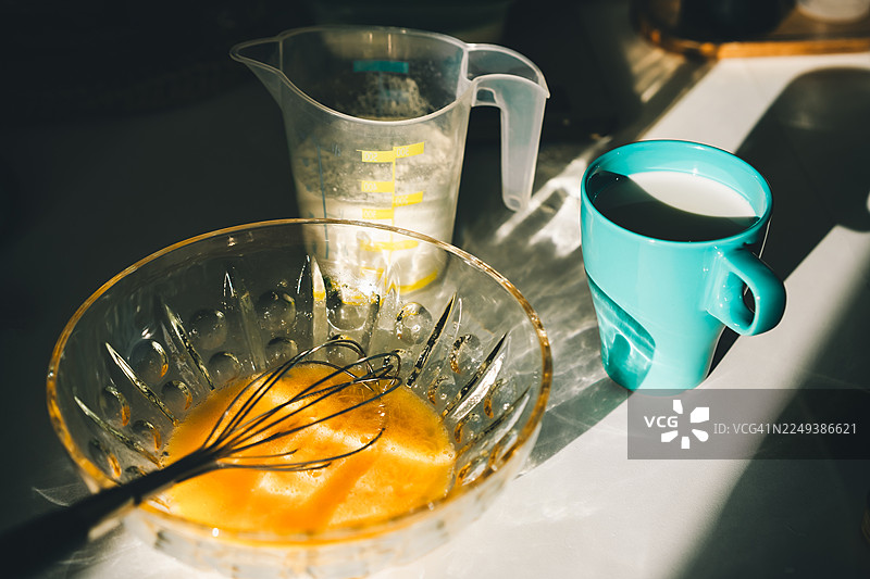 在阳光明媚的厨房台面上打鸡蛋并准备食材，包括一个玻璃碗、一个牛奶杯和一个量杯。图片素材