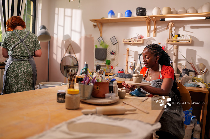 一位年轻女子在光线充足的手工坊里，使用陶轮勤奋地工作，塑造着陶罐，展现了创造力和工艺精神。图片素材