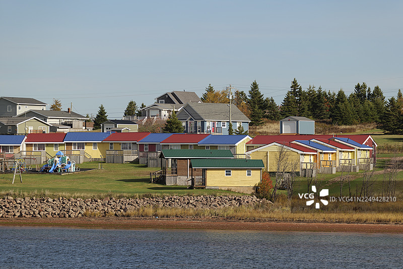 色彩缤纷的房屋和小屋沿着绿草如茵的岸边排列，旁边是一片水域，天空湛蓝清澈。图片素材