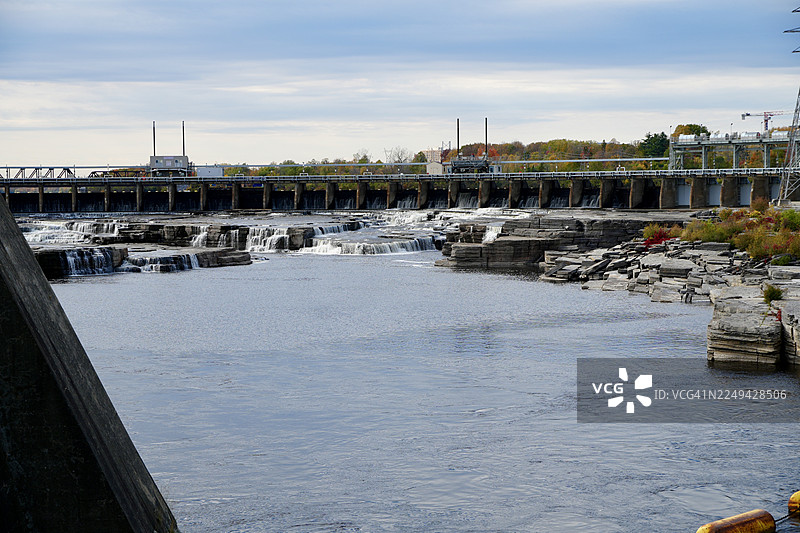 阴天下的水电站和瀑布的风景，河岸两旁展示着秋天的落叶图片素材