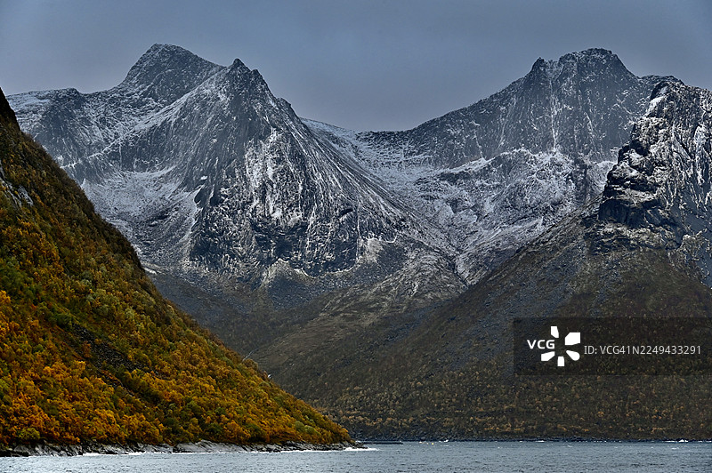 挪威特罗姆郡森雅岛赫斯泰斯科峰，秋季色彩斑斓的树木环绕着湖泊，远处是白雪皑皑的壮丽山峦，天空布满云彩，构成了一幅戏剧性的风景画图片素材