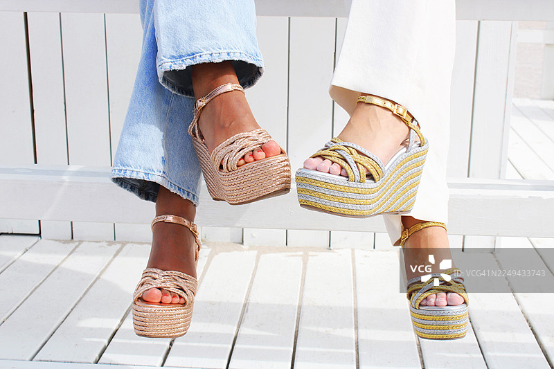 两位女士穿着时尚的坡跟草编鞋，站在白色的木制甲板上，展示着夏日时尚鞋履图片素材