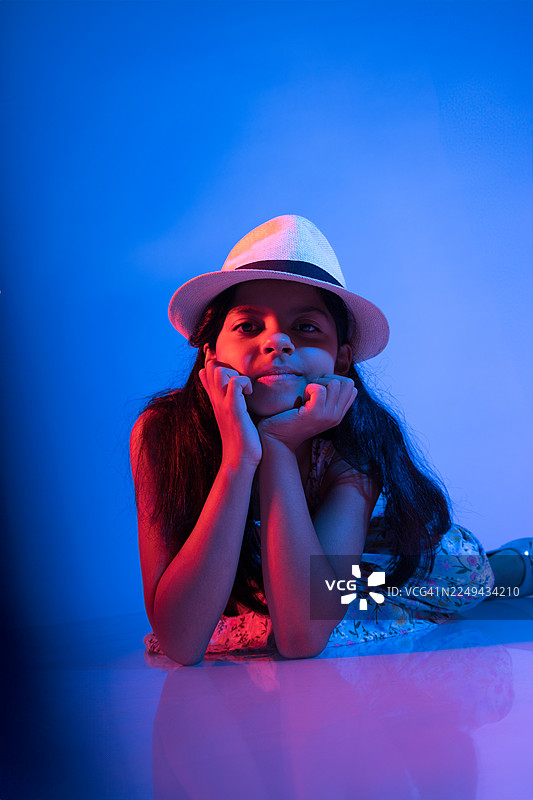 一个戴着帽子的年轻女孩的肖像，她趴着，双手放在下巴下面，背景是鲜艳的蓝色。图片素材