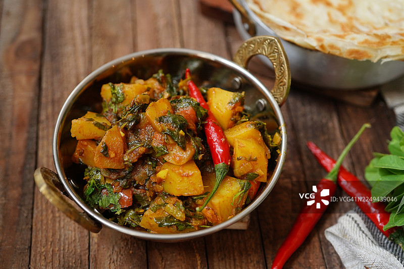 俯视角度拍摄的美味土豆菠菜咖喱，装在传统的金属碗中，用红辣椒装饰，放在乡村木桌上，背景有扁平面包。图片素材