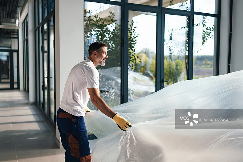 身穿白色T恤的男子在正在施工的现代化办公室里展开大块塑料布图片素材
