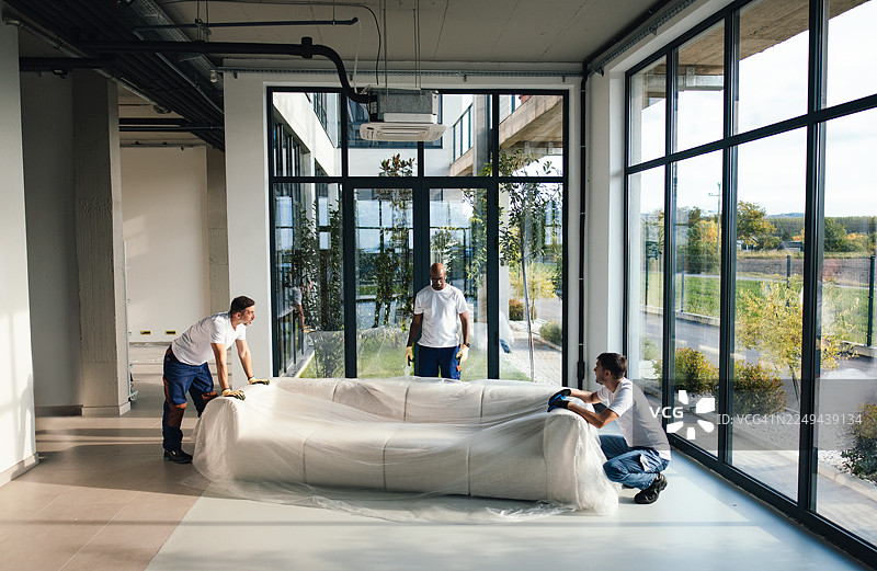 团队工作人员在搬家过程中，在现代办公空间内拆箱并包裹沙发图片素材