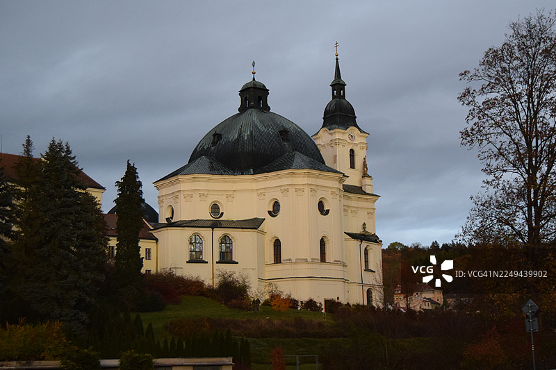 从低角度看一座历史悠久的教堂，它拥有一个大圆顶和尖塔，背景是戏剧性的多云天空，周围环绕着秋天的树木，捷克共和国图片素材