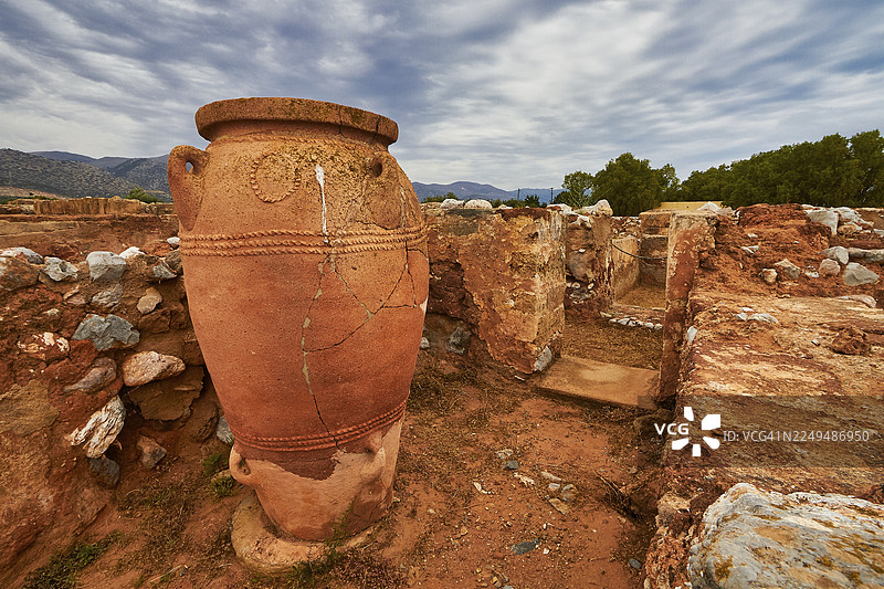 一座大型米诺斯陶罐矗立在废墟之中，天空多云，周围环绕着石头和大地色调，这里是联合国教科文组织世界遗产——克里特岛马利亚米诺斯宫遗址。图片素材