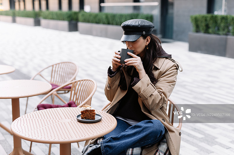 年轻的棕发女子穿着休闲服，坐在城市街道旁的户外咖啡馆的桌子旁喝咖啡，展现都市生活和休闲理念图片素材