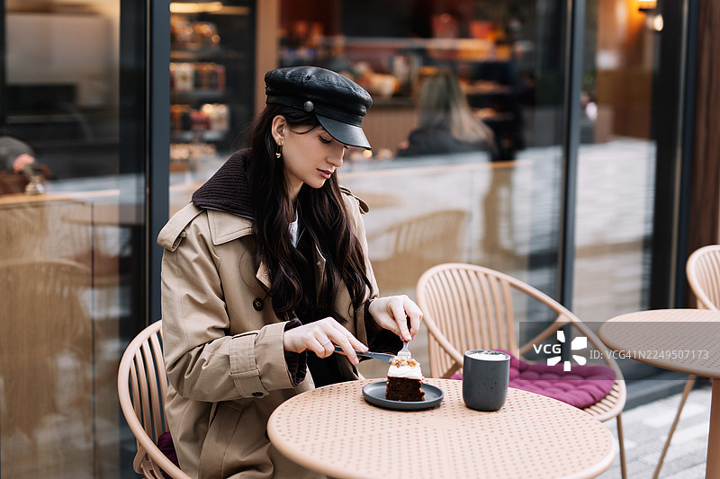 年轻的棕发女子身着休闲服饰和头饰，在街边公共场所的户外咖啡馆桌旁，一边吃着甜点一边喝着咖啡。图片素材