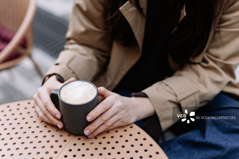 一位年轻的棕发女性，身穿休闲服饰，坐在城市街道旁的户外咖啡馆桌子旁喝咖啡，体现都市生活方式和休闲概念图片素材