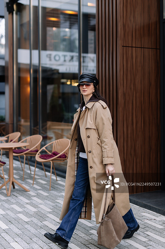 戴着太阳镜和帽子的年轻时尚女子，身着时髦休闲服装，手持绒面革包，行走在城市街道上。都市时尚。叠穿风衣造型概念。图片素材