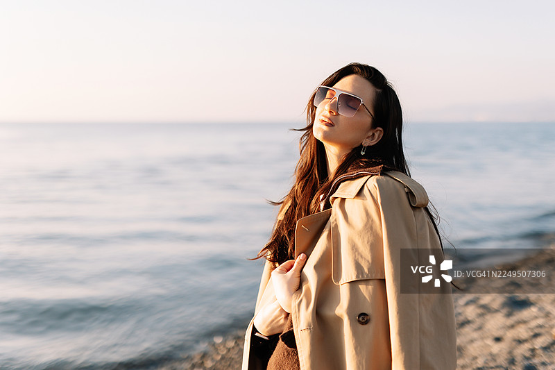 一位穿着毛衣和外套的美丽棕发女子，在阳光明媚的淡季天气里，独自一人在大自然海边漫步。心理健康与独自旅行的概念图片素材