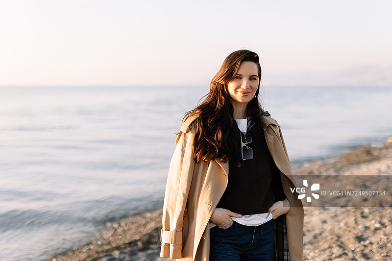 一位美丽的棕发女子，身穿毛衣和外套，独自一人在阳光明媚的淡季，在大自然的海边漫步。心理健康和独自旅行的概念。图片素材