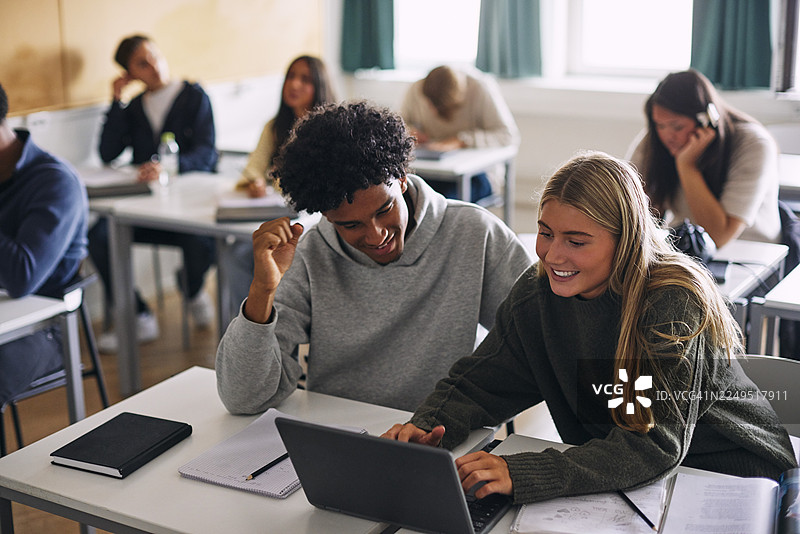 高中教室里，一位微笑的少女正和她的男性朋友一起学习，并使用笔记本电脑。图片素材