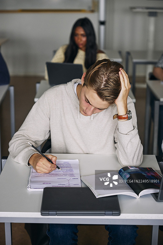 俯视角度，年轻男子在教室里倚着胳膊在书本上写作图片素材
