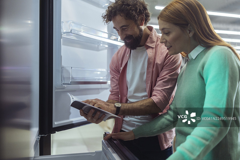 一家电子产品商店里，一对无忧无虑的恩爱夫妻从侧面查看展出的冰箱图片素材