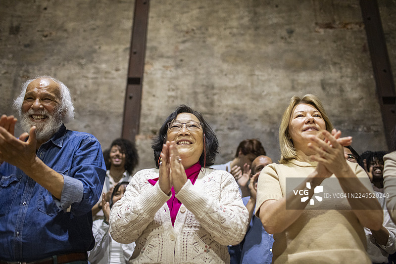 从低角度看，不同种族的老年男女观众在剧院起立鼓掌。图片素材