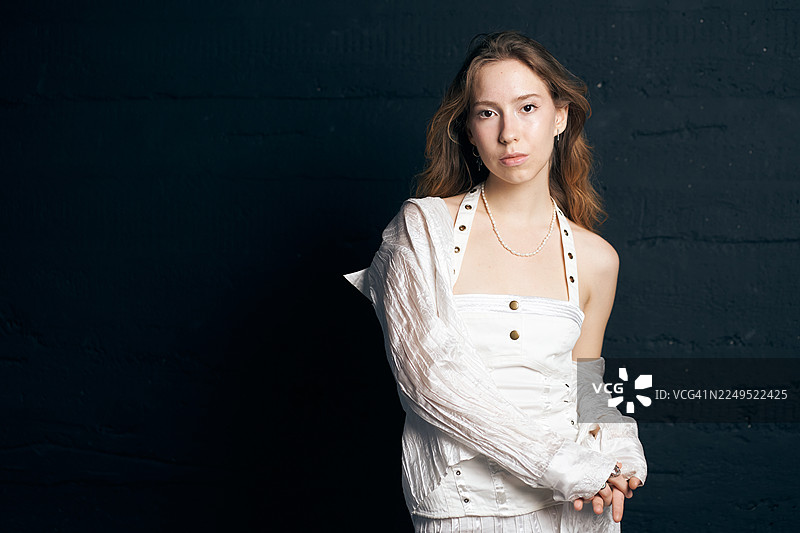 身着白色服装的年轻女子在工作室风格的人像摄影中，摆姿势面对深色纹理墙壁。2026年度代表色，云舞者。图片素材