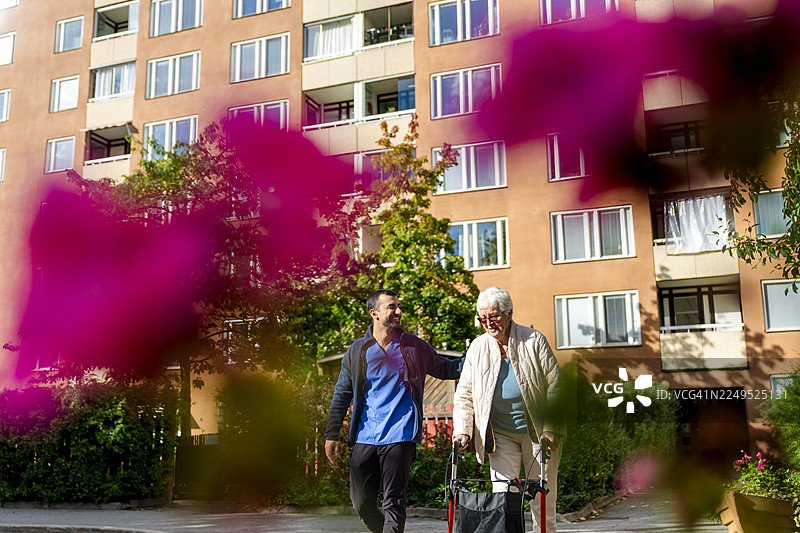 晴天在公园里，一名男性医护人员陪同使用助行器的老年女性行走，低角度拍摄。图片素材