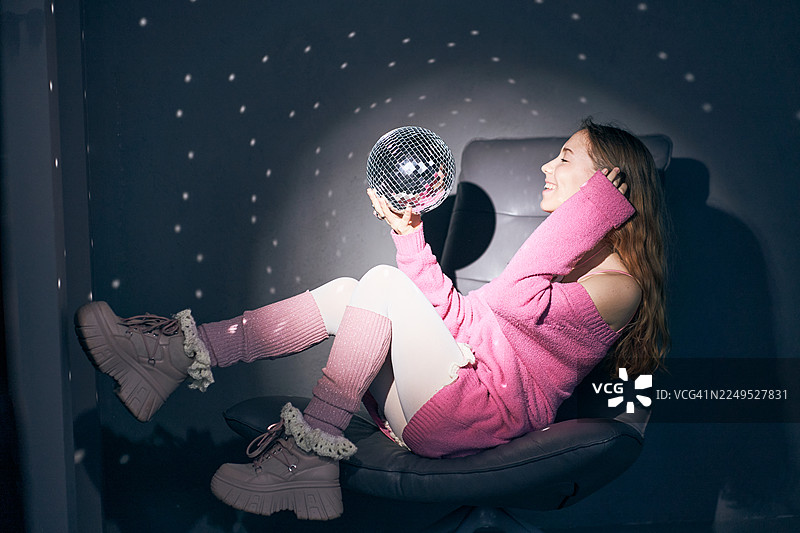 身穿粉色毛衣的年轻女子在昏暗的房间里放松，房间里有一个迪斯科球和柔和的反射光。图片素材