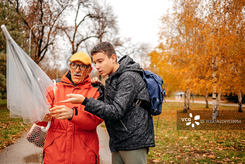 在雨天，一位老人与他的十几岁的孙子在公园见面，并撑开雨伞。图片素材