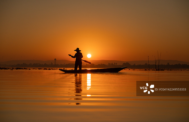 剪影渔夫站在木船上，在平静的水面上撒网，展现了传统的捕鱼方式和场景的自然和谐。图片素材