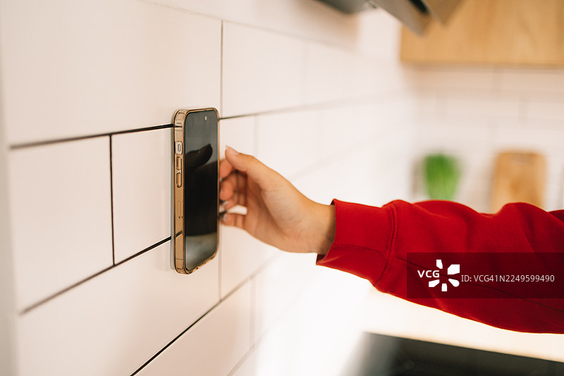 一个穿着红袖子的人伸手去拿固定在白色瓷砖墙上的智能手机，手机用吸盘手机壳固定图片素材