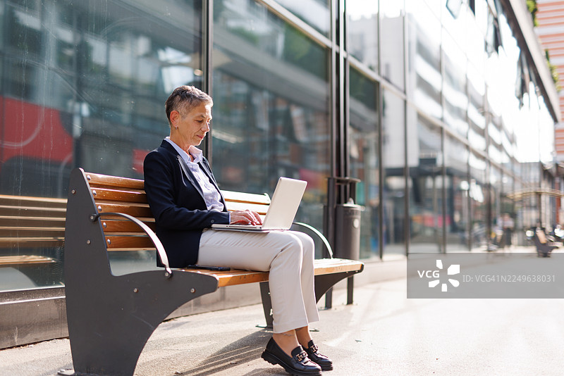 一位年长的女商人在城市户外使用现代笔记本电脑工作，敲击键盘并使用科技产品。图片素材
