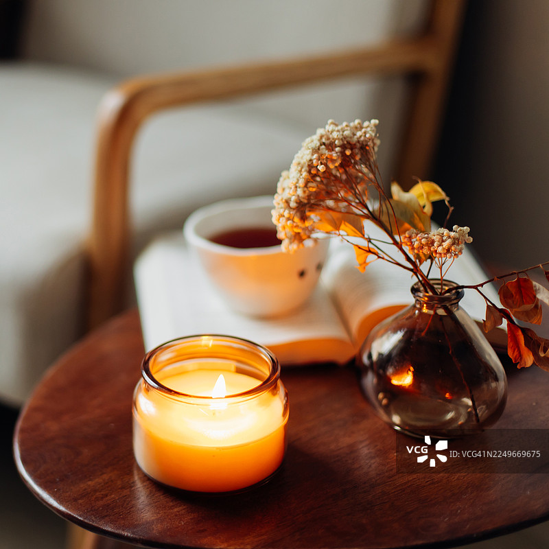 一个舒适的居家阅读角，配有木制扶手椅和一个放着茶杯、打开的书籍和眼镜的小圆边桌，以及一支点亮的蜡烛。温暖的自然光和柔和的大地色调营造出平静放松的氛围。图片素材