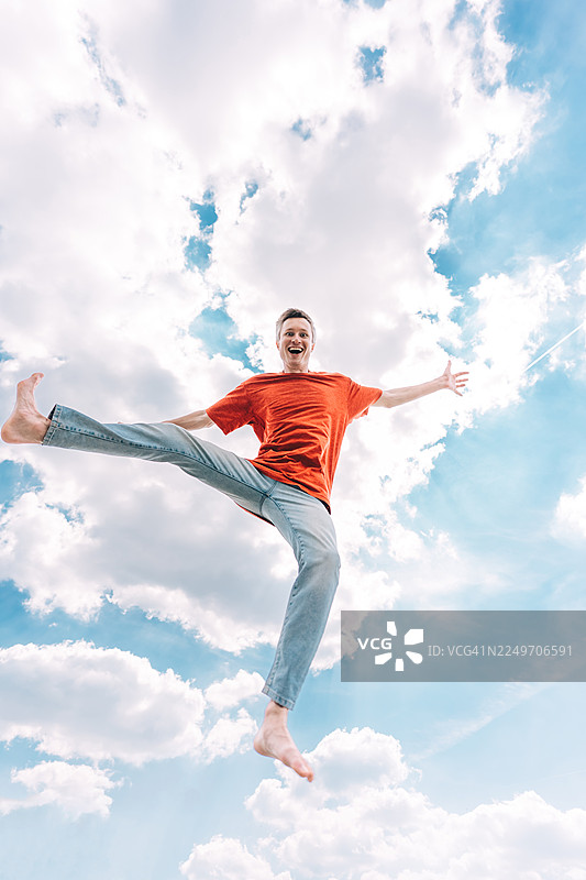 一个穿着红色T恤的年轻男子在天空背景下快乐地跳跃，全身镜头。图片素材