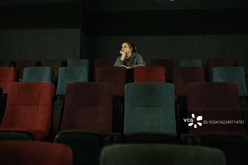 一位沉思的女子独自坐在昏暗的电影院里，周围是彩色的座位，她若有所思。图片素材