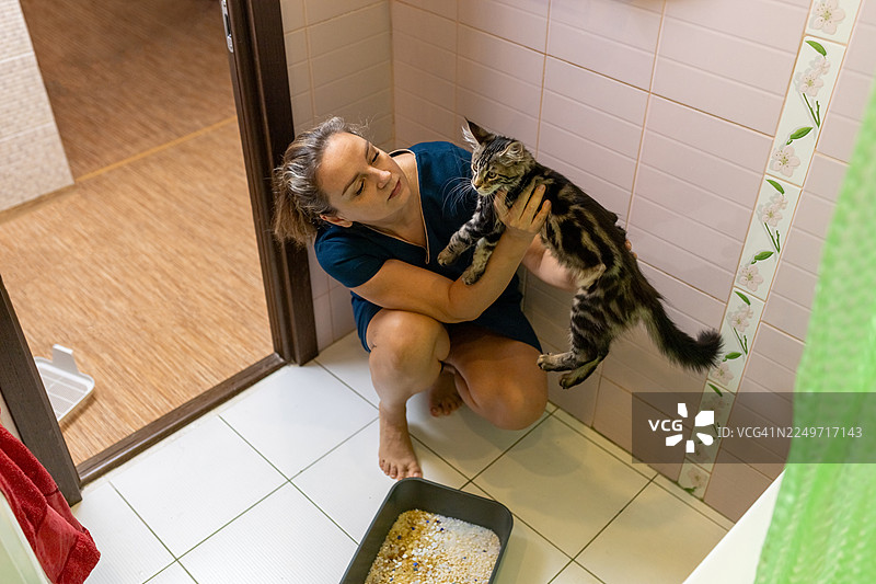 一位女士在浴室里轻轻抱着一只猫，旁边正在清理猫砂盆，分享着充满关爱的时刻图片素材