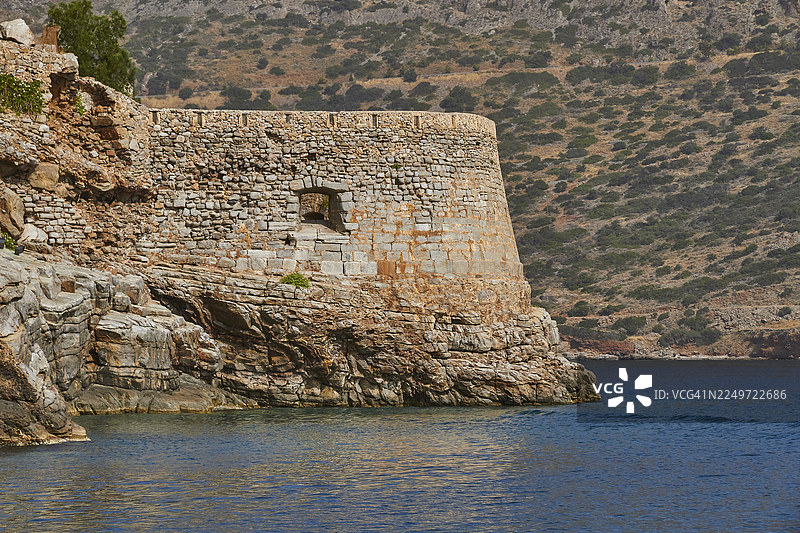 希腊克里特岛东部米拉贝罗湾埃卢达湾的斯皮纳隆加勒珀岛威尼斯海堡的岩石海岸历史要塞墙，天空湛蓝，海水平静图片素材