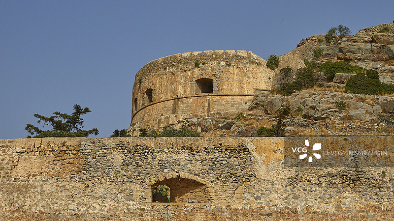 圆形石塔矗立在岩石地面上,四周环绕着古老的城墙和植被,这是威尼斯海滨要塞,位于麻风岛、斯皮纳隆加、埃卢达湾、米拉贝洛湾,属于希腊克里特岛东部,希腊群岛,希腊图片素材