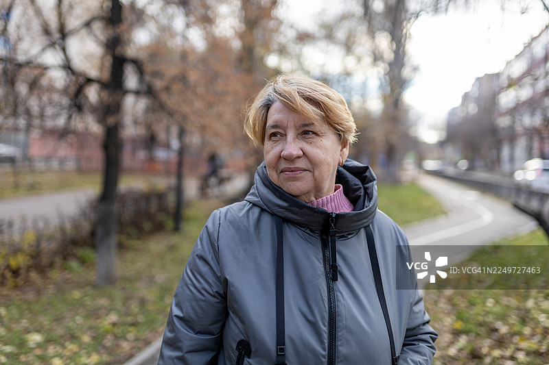 一位穿着灰色夹克的老年女性在秋日宁静的公园里，目光转向一侧图片素材