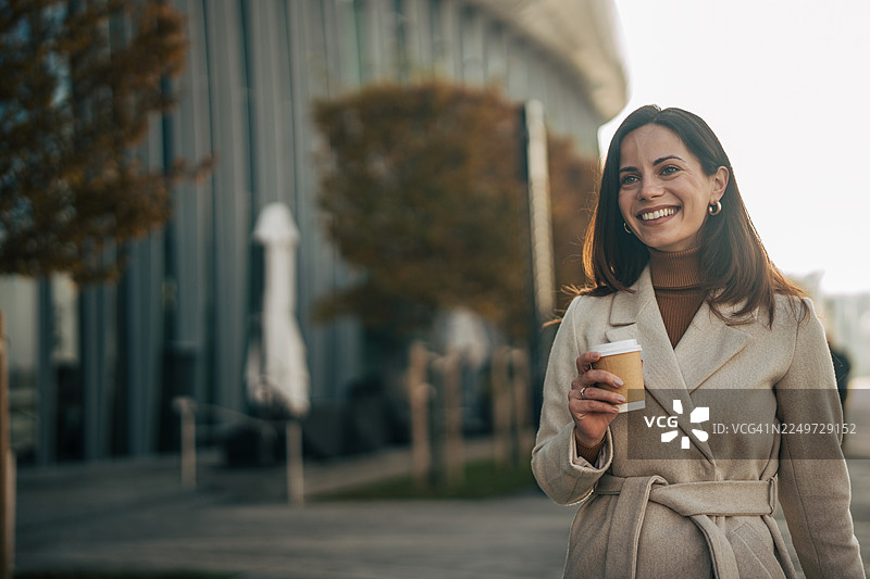 快乐的女商人一手拿着咖啡，一手走在城市里图片素材