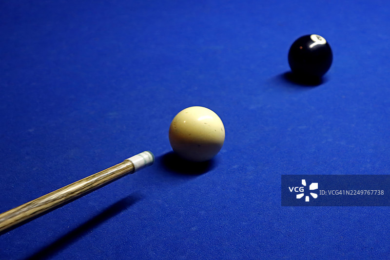 特写镜头，低饱和度戏剧性光线下的白色母球和黑色8号球，放置在鲜艳的蓝色斯诺克或台球桌上，木制球杆的杆头瞄准母球附近。图片素材