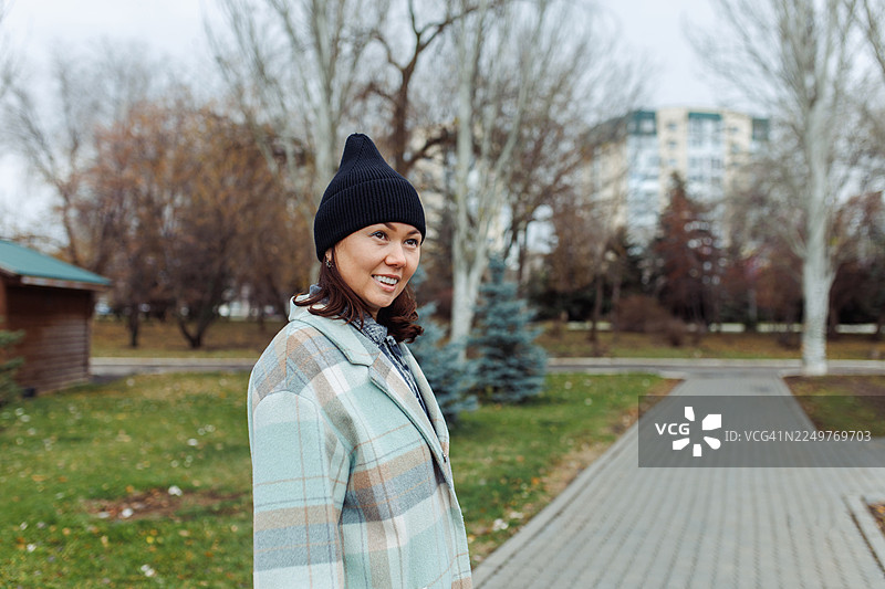 秋日公园里，一位戴着黑色绒线帽、身穿格子外套的女士面带微笑图片素材
