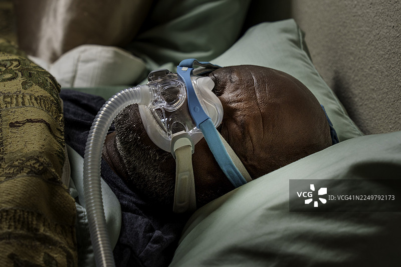 一位年长的非裔美国男性在室内睡觉，佩戴着睡眠呼吸暂停面罩和呼吸设备。图片素材