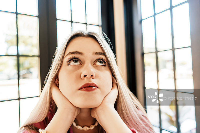 年轻女子在明亮的窗边空间向上凝视，双手托着下巴图片素材