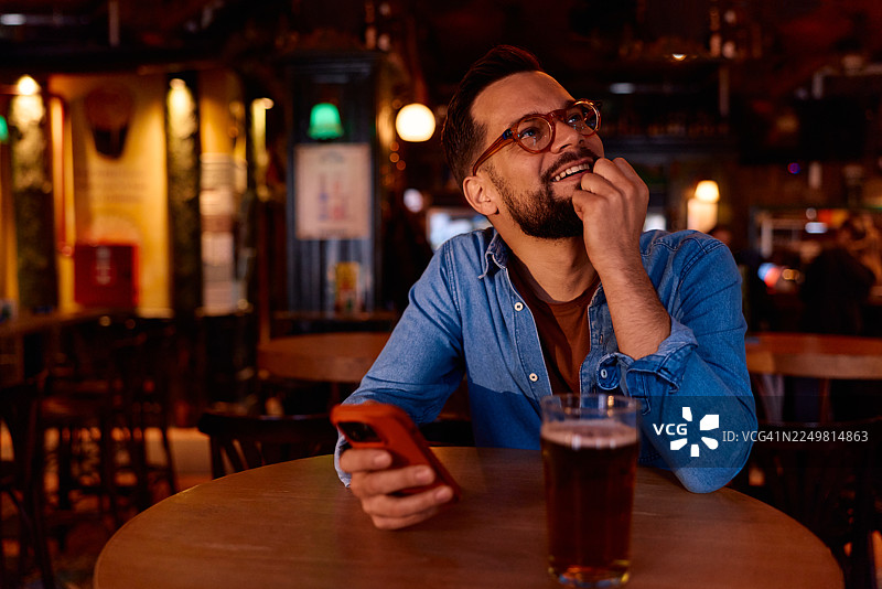 男人在酒吧里边享受饮品边看手机，脸上带着微笑看向别处图片素材