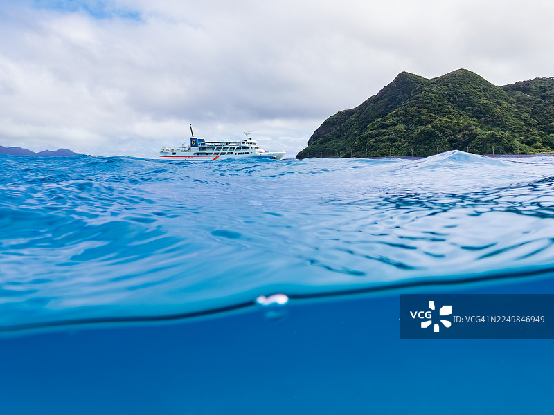 从美妙的珊瑚礁水面拍摄的渡嘉敷岛渡轮“座间味号”和古座间岛的美丽半水景。一群热带鱼。美丽的珊瑚礁和游泳的人们。阿嘉海滩，阿嘉岛，冲绳，渡嘉敷图片素材